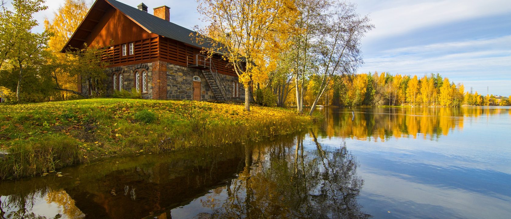 Syksyinen maisema, jossa kellastuneita puita ja mökki järven rannalla. Sininen taivas.